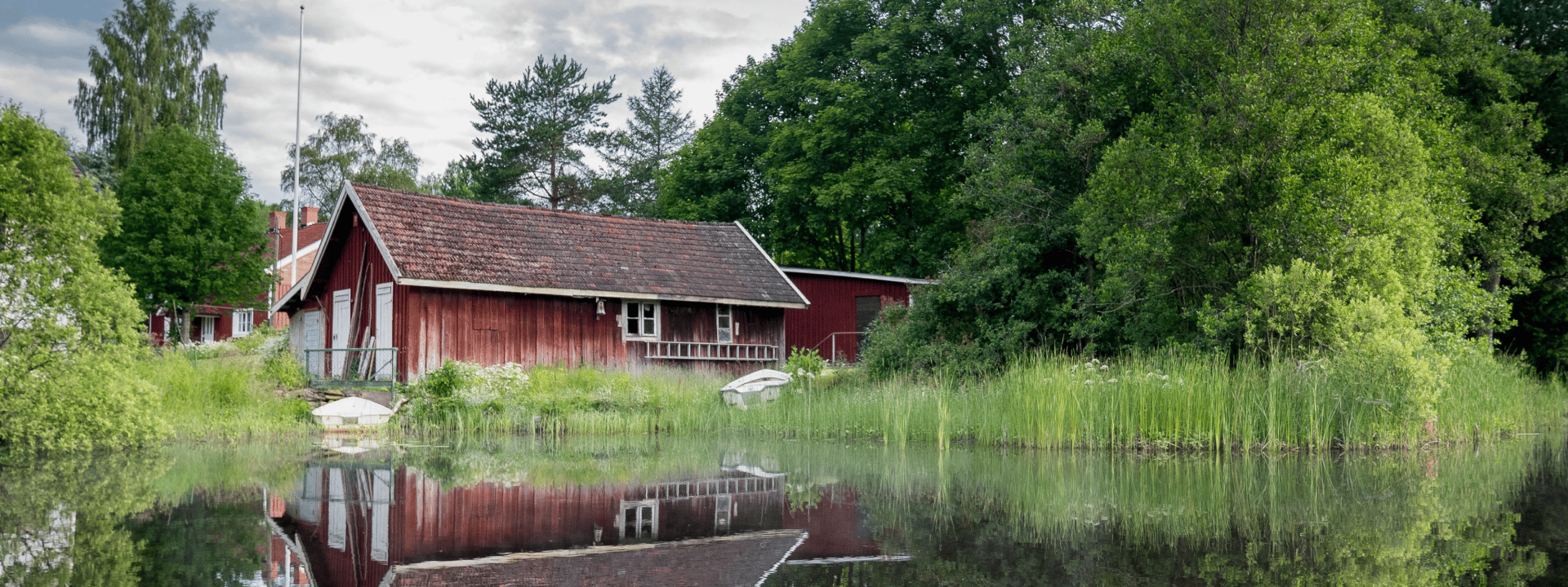 Drybox Ett rött trähus med sluttande tak ligger vid en lugn sjö, omgivet av frodiga gröna träd och buskar. Tack vare vindsavfuktare förblir vinden torr året runt. En liten vit båt vilar på stranden, reflekterad i det stilla vattnet.