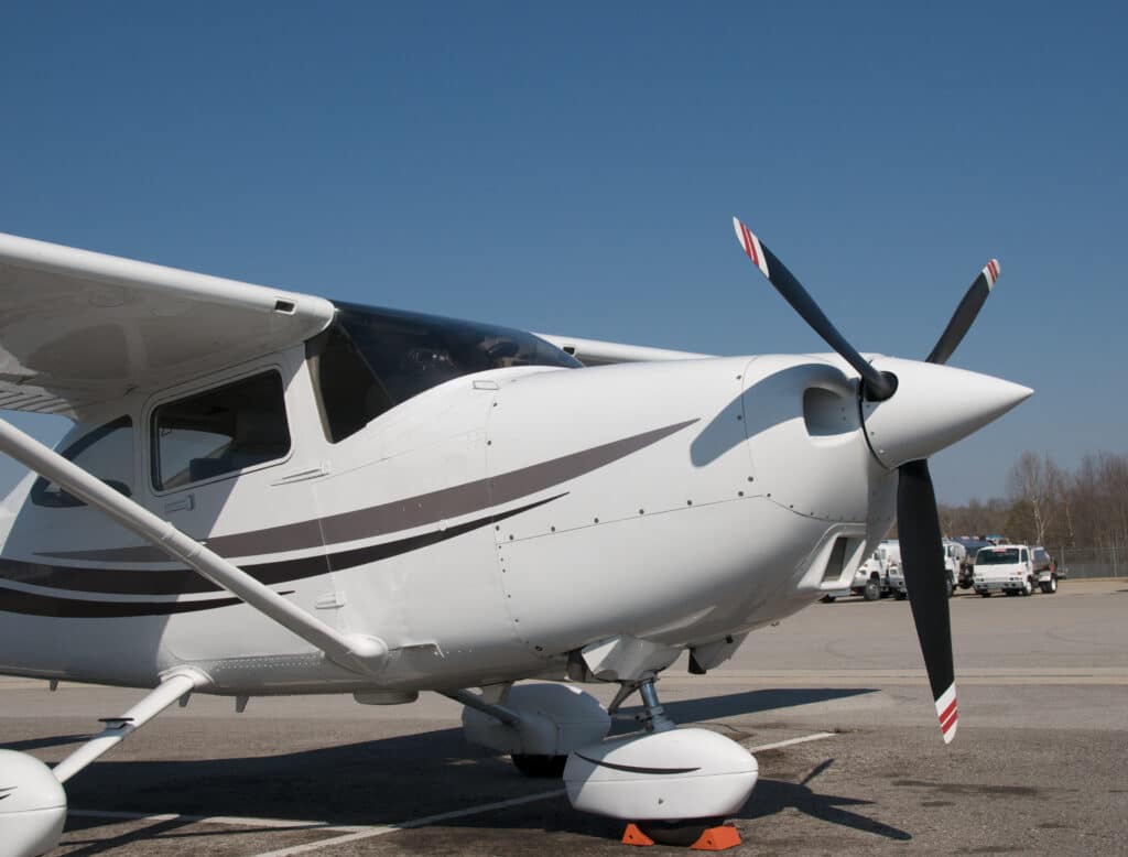 Drybox Ett litet vitt enmotorigt flygplan med bruna ränder står parkerat på en asfaltsbana under en klarblå himmel. Propellern står stilla, orange hjulklossar säkrar noshjulet och en Avfuktare-skåpbil syns bland andra fordon i bakgrunden.