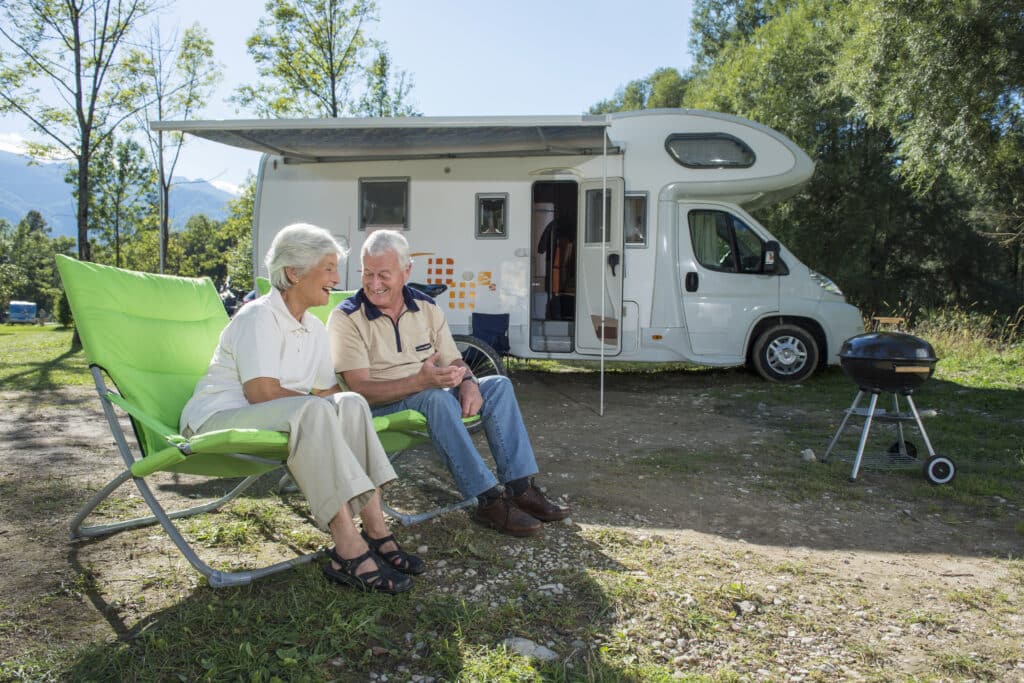 Drybox Ett äldre par sitter leende på gröna campingstolar utanför sin husbil och njuter av en drink och ett samtal bredvid en grill och en bärbar Avfuktare på den soliga, skogsklädda campingplatsen.